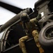 A military aviator or crew member in full combat gear and flight helmet is climbing onto or inspecting the exterior of a military helicopter, likely a UH-60 Black Hawk. The individual is wearing multicam/OCP camouflage uniform and appears to be conducting a pre-flight or post-flight inspection. The image is taken from a low angle, emphasizing the scale of the aircraft and the crew member's activity.
