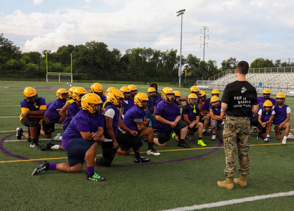 A U.S. Marine in camouflage utilities and a black t-shirt reading 'Pain is Weakness Leaving the Body' addresses a high school football team on a practice field. The players are wearing purple jerseys and gold helmets, kneeling or sitting attentively on the turf. This appears to be a Marine Corps outreach or mentorship event at a high school athletic facility.