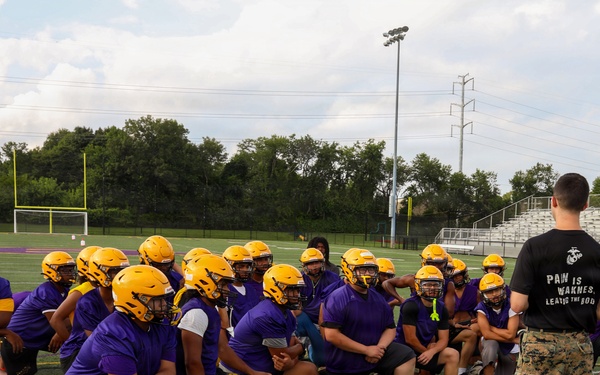 A U.S. Marine in camouflage utilities and a black t-shirt reading 'Pain is Weakness Leaving the Body' addresses a high school football team on a practice field. The players are wearing purple jerseys and gold helmets, kneeling or sitting attentively on the turf. This appears to be a Marine Corps outreach or mentorship event at a high school athletic facility.