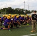 A U.S. Marine in camouflage utilities and a black t-shirt reading 'Pain is Weakness Leaving the Body' addresses a high school football team on a practice field. The players are wearing purple jerseys and gold helmets, kneeling or sitting attentively on the turf. This appears to be a Marine Corps outreach or mentorship event at a high school athletic facility.