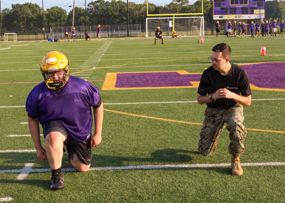 A U.S. Marine in camouflage utilities is coaching or instructing a high school football player wearing a purple jersey and gold helmet during a practice session on a turf field. The field features purple and gold team colors, consistent with a school's branding. Multiple other players and coaches are visible in the background engaged in various drills.