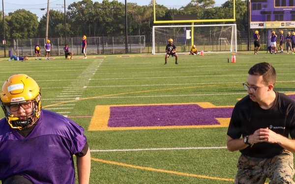A U.S. Marine in camouflage utilities is coaching or instructing a high school football player wearing a purple jersey and gold helmet during a practice session on a turf field. The field features purple and gold team colors, consistent with a school's branding. Multiple other players and coaches are visible in the background engaged in various drills.