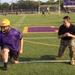 A U.S. Marine in camouflage utilities is coaching or instructing a high school football player wearing a purple jersey and gold helmet during a practice session on a turf field. The field features purple and gold team colors, consistent with a school's branding. Multiple other players and coaches are visible in the background engaged in various drills.