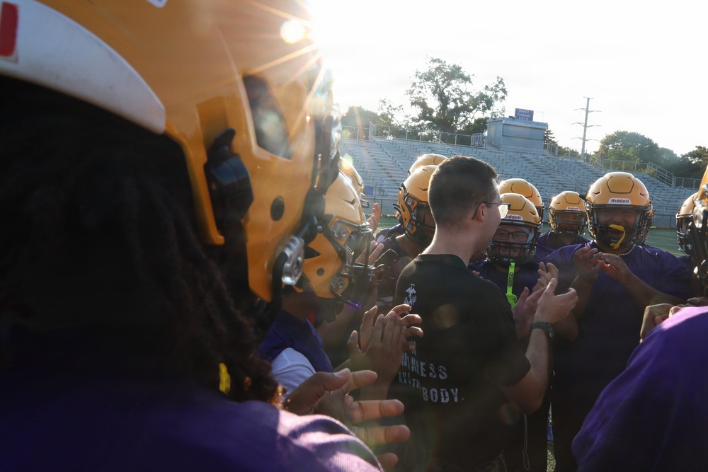 A football team huddle or pre-game ritual is taking place on a field, with players wearing gold/yellow helmets and purple uniforms exchanging high-fives or handshakes. A coach or staff member in a black t-shirt stands at the center of the group. The image is backlit by strong sunlight, creating lens flare and a dramatic atmosphere.
