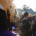A football team huddle or pre-game ritual is taking place on a field, with players wearing gold/yellow helmets and purple uniforms exchanging high-fives or handshakes. A coach or staff member in a black t-shirt stands at the center of the group. The image is backlit by strong sunlight, creating lens flare and a dramatic atmosphere.
