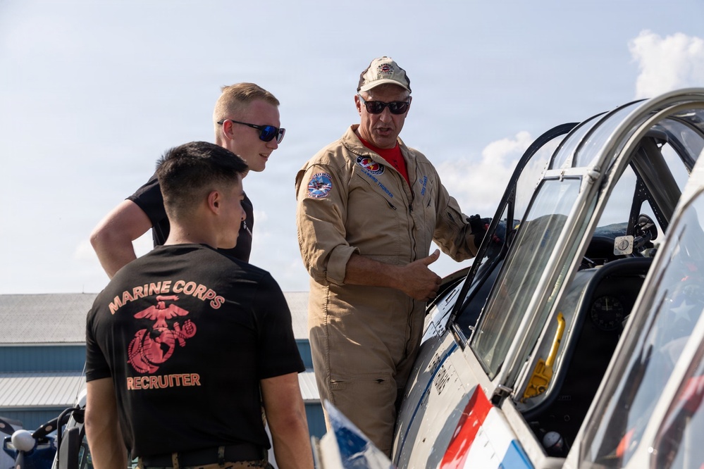 A pilot in a tan flight suit is explaining or demonstrating features of a military jet aircraft to two young men at what appears to be an airshow or airfield. One of the young men is wearing a black 'Marine Corps Recruiter' t-shirt, suggesting a military recruitment or outreach context. The scene takes place outdoors on a sunny day near a hangar.