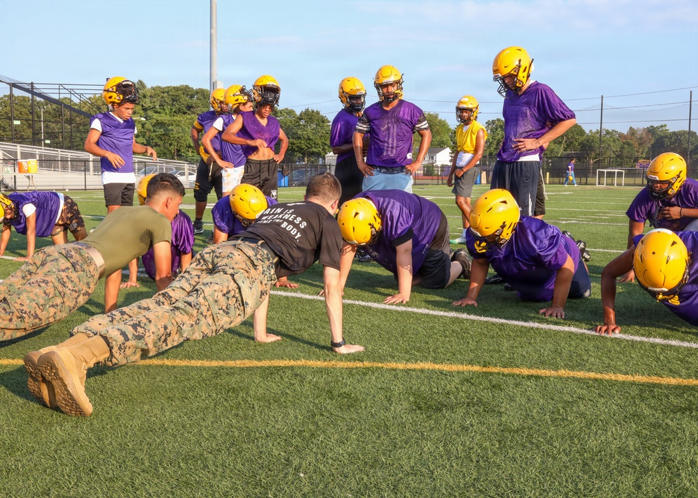 A U.S. Marine in camouflage uniform is demonstrating push-ups to a youth or high school football team on a practice field. Several players in purple jerseys and gold helmets are performing push-ups alongside the Marine, while others stand and observe. The scene appears to be a military outreach or community engagement event during a football practice session.