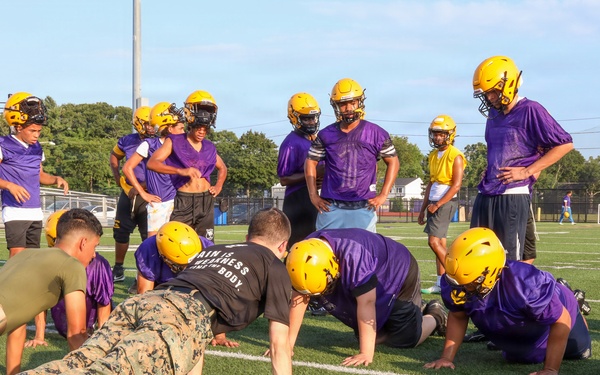 A U.S. Marine in camouflage uniform is demonstrating push-ups to a youth or high school football team on a practice field. Several players in purple jerseys and gold helmets are performing push-ups alongside the Marine, while others stand and observe. The scene appears to be a military outreach or community engagement event during a football practice session.