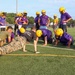 A U.S. Marine in camouflage uniform is demonstrating push-ups to a youth or high school football team on a practice field. Several players in purple jerseys and gold helmets are performing push-ups alongside the Marine, while others stand and observe. The scene appears to be a military outreach or community engagement event during a football practice session.