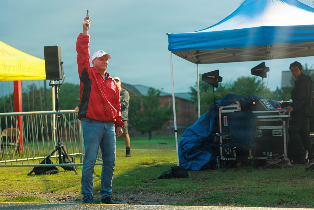 A man in a red jacket and white cap holds a starting pistol raised above his head at what appears to be an outdoor race or sporting event. The setting includes event tents, audio/PA equipment, and crowd barriers, suggesting an organized public competition. The overcast sky and wet ground indicate rainy or recently rainy conditions.