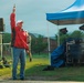 A man in a red jacket and white cap holds a starting pistol raised above his head at what appears to be an outdoor race or sporting event. The setting includes event tents, audio/PA equipment, and crowd barriers, suggesting an organized public competition. The overcast sky and wet ground indicate rainy or recently rainy conditions.