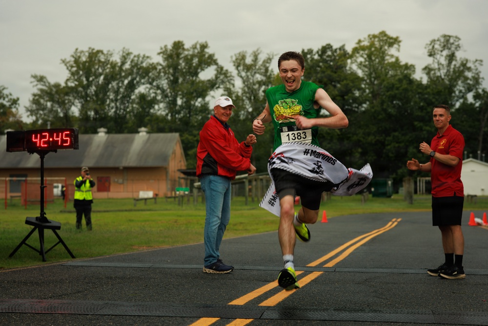 A runner wearing bib number 1383 crosses the finish line of a road race with an expression of triumph and joy. The digital clock in the background displays a finishing time of 42:45. Race officials and spectators are present on both sides of the finish line, with one official extending a hand toward the winner.