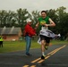 A runner wearing bib number 1383 crosses the finish line of a road race with an expression of triumph and joy. The digital clock in the background displays a finishing time of 42:45. Race officials and spectators are present on both sides of the finish line, with one official extending a hand toward the winner.