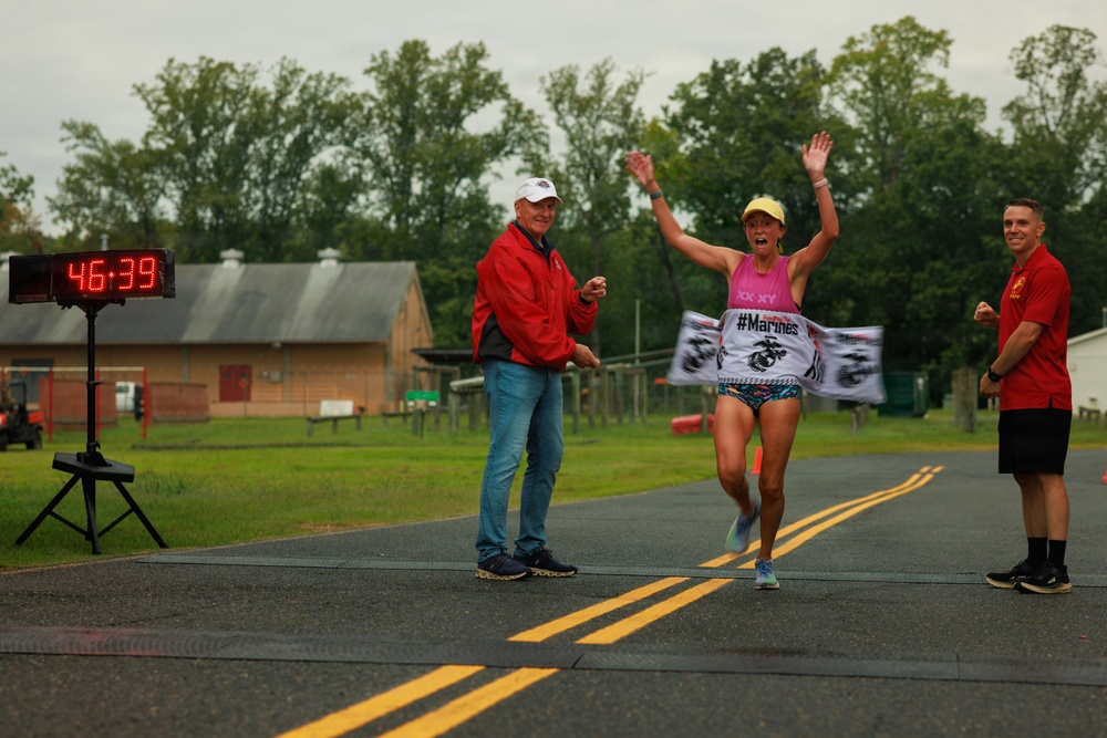 A female runner is crossing the finish line of a race with arms raised in celebration, breaking through a '#Marines' finish line banner. Two officials in red jackets are present at the finish line, and a digital race clock displays a time of 46:39. The event appears to be a military-affiliated road race held on a paved road in a rural or base setting.