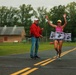 A female runner is crossing the finish line of a race with arms raised in celebration, breaking through a '#Marines' finish line banner. Two officials in red jackets are present at the finish line, and a digital race clock displays a time of 46:39. The event appears to be a military-affiliated road race held on a paved road in a rural or base setting.
