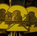 The image shows a close-up of a military medal belonging to the Second Recruit Training Battalion of the United States Marine Corps. The medal features a bronze finish with a yellow enamel background shaped as the number '2', adorned with crossed swords, a drill instructor's campaign cover (hat), and the USMC Eagle, Globe, and Anchor emblem. Multiple identical medals are visible in the background, suggesting a batch prepared for a graduation or award ceremony.
