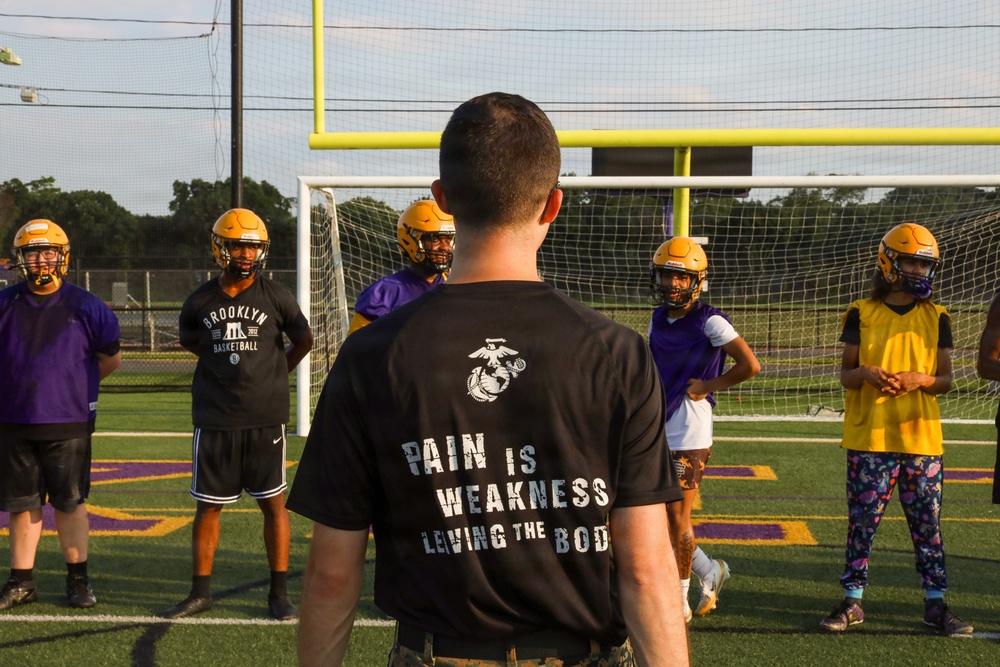 A person wearing a black U.S. Marine Corps t-shirt reading 'Pain is Weakness Leaving the Body' stands with their back to the camera, addressing a group of young football players wearing gold helmets and purple/yellow jerseys. The setting is an outdoor athletic field with yellow goal posts and a soccer net visible in the background. This appears to be a football practice or training session, likely with a military recruiter or coach leading the group.