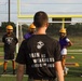 A person wearing a black U.S. Marine Corps t-shirt reading 'Pain is Weakness Leaving the Body' stands with their back to the camera, addressing a group of young football players wearing gold helmets and purple/yellow jerseys. The setting is an outdoor athletic field with yellow goal posts and a soccer net visible in the background. This appears to be a football practice or training session, likely with a military recruiter or coach leading the group.