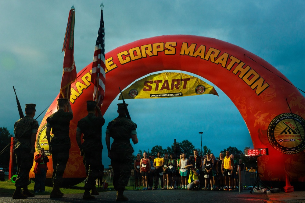 The image shows the start line of the Marine Corps Marathon, with a large red inflatable arch bearing the event name and a yellow 'START' banner with the hashtag '#RUNWITHTHEMARINES'. A military color guard stands at attention on the left side holding flags, including the U.S. flag and a Marine Corps flag, while a large group of runners is assembled beneath the arch awaiting the race start. A digital race clock displays '6:25:16' in the background, suggesting an early morning pre-race moment.