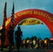 The image shows the start line of the Marine Corps Marathon, with a large red inflatable arch bearing the event name and a yellow 'START' banner with the hashtag '#RUNWITHTHEMARINES'. A military color guard stands at attention on the left side holding flags, including the U.S. flag and a Marine Corps flag, while a large group of runners is assembled beneath the arch awaiting the race start. A digital race clock displays '6:25:16' in the background, suggesting an early morning pre-race moment.