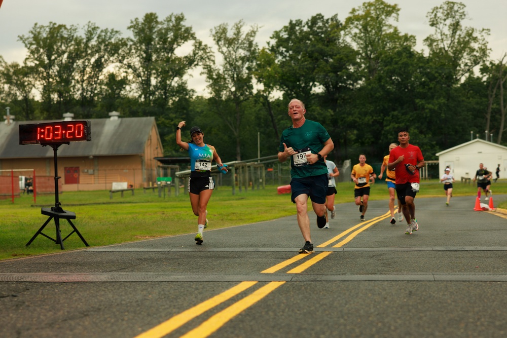A road race is in progress, with multiple runners approaching what appears to be the finish line area. The race clock displays 1:03:20, indicating elapsed time. A woman wearing bib number 142 is visibly celebrating with a raised fist, while a man wearing bib number 197 leads the foreground group.