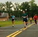 A road race is in progress, with multiple runners approaching what appears to be the finish line area. The race clock displays 1:03:20, indicating elapsed time. A woman wearing bib number 142 is visibly celebrating with a raised fist, while a man wearing bib number 197 leads the foreground group.
