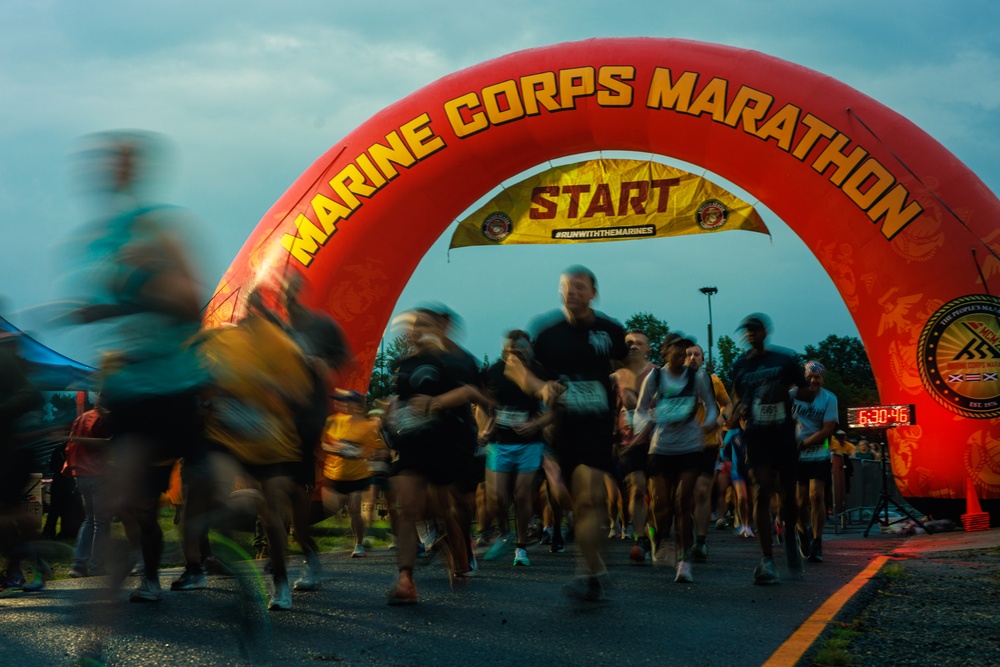 Runners are crossing the start line of the Marine Corps Marathon beneath a large red inflatable arch. The image was taken at dawn or early morning, capturing the energy and motion blur of participants beginning the race. A digital clock on the right displays a time of 6:30:46, indicating the race has just begun.