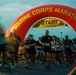 Runners are crossing the start line of the Marine Corps Marathon beneath a large red inflatable arch. The image was taken at dawn or early morning, capturing the energy and motion blur of participants beginning the race. A digital clock on the right displays a time of 6:30:46, indicating the race has just begun.