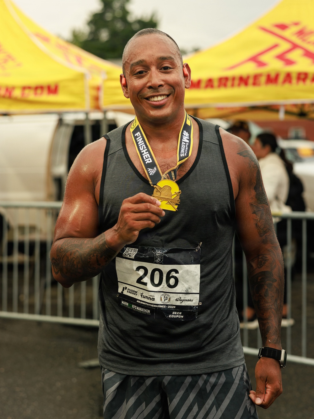 A muscular man with tattoos on both arms is posing proudly after completing a marathon race. He is wearing bib number 206 and displaying a gold finisher's medal around his neck. The event appears to be the 'Run with the Marines' marathon, based on visible text on the bib and background banners.