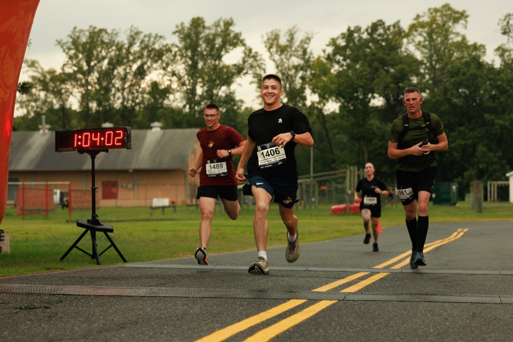 A road race is in progress, with multiple runners approaching what appears to be a finish line area. The lead runner, wearing bib number 1406 and a black shirt, is smiling broadly as he nears the finish. A digital race clock displays a time of 1:04:02, and several other competitors with visible bib numbers are close behind.