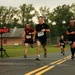 A road race is in progress, with multiple runners approaching what appears to be a finish line area. The lead runner, wearing bib number 1406 and a black shirt, is smiling broadly as he nears the finish. A digital race clock displays a time of 1:04:02, and several other competitors with visible bib numbers are close behind.