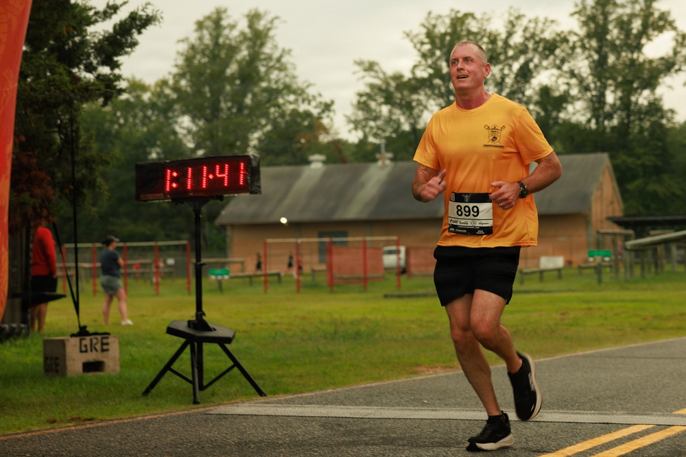 A male runner wearing bib number 899 and an orange t-shirt is captured mid-stride during a road race on a wet, overcast day. A digital race clock in the background displays a time of 1:11:41. The setting appears to be a park or fairground with wooden buildings and green grass visible in the background.