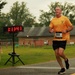 A male runner wearing bib number 899 and an orange t-shirt is captured mid-stride during a road race on a wet, overcast day. A digital race clock in the background displays a time of 1:11:41. The setting appears to be a park or fairground with wooden buildings and green grass visible in the background.