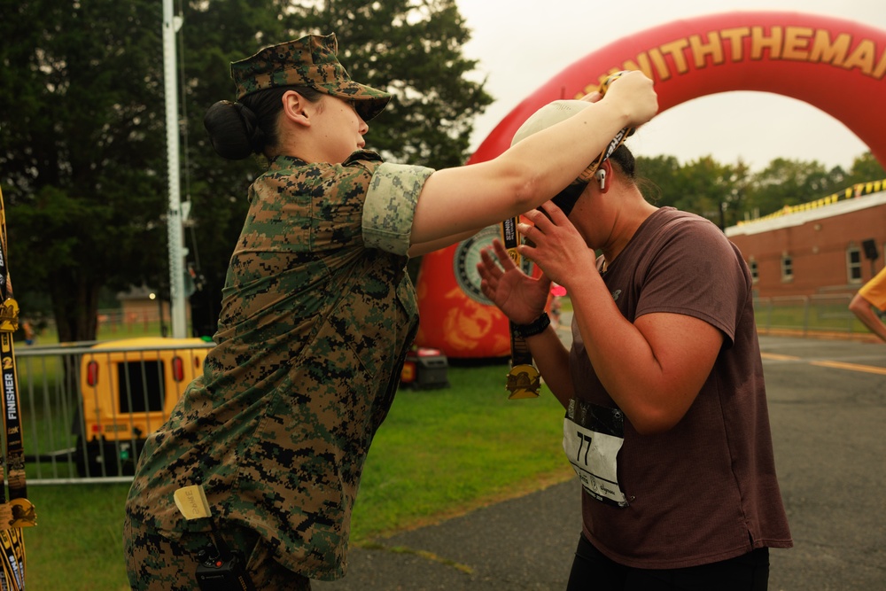 A U.S. Marine in digital camouflage uniform is placing a finisher medal over the head of a race participant wearing bib number 77. The scene takes place at what appears to be a Marine Corps-sponsored running event, with a large red inflatable arch visible in the background bearing partial text 'WITHTHEMA' (likely 'Run With The Marines'). The runner is bowing their head to receive the medal at the finish area.
