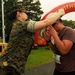 A U.S. Marine in digital camouflage uniform is placing a finisher medal over the head of a race participant wearing bib number 77. The scene takes place at what appears to be a Marine Corps-sponsored running event, with a large red inflatable arch visible in the background bearing partial text 'WITHTHEMA' (likely 'Run With The Marines'). The runner is bowing their head to receive the medal at the finish area.