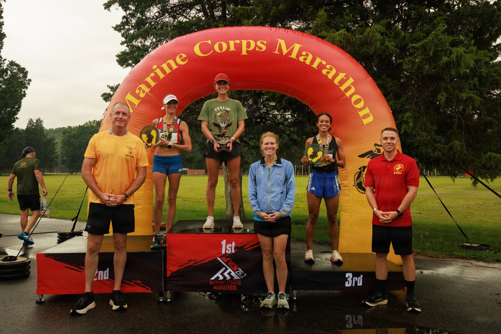 A podium ceremony at the Marine Corps Marathon featuring three female finishers standing on 1st, 2nd, and 3rd place platforms beneath a large inflatable arch. The event is taking place outdoors in wet, overcast conditions, with two officials flanking the podium. The first-place finisher holds a trophy while the 2nd and 3rd place finishers hold medals/awards.