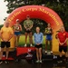 A podium ceremony at the Marine Corps Marathon featuring three female finishers standing on 1st, 2nd, and 3rd place platforms beneath a large inflatable arch. The event is taking place outdoors in wet, overcast conditions, with two officials flanking the podium. The first-place finisher holds a trophy while the 2nd and 3rd place finishers hold medals/awards.