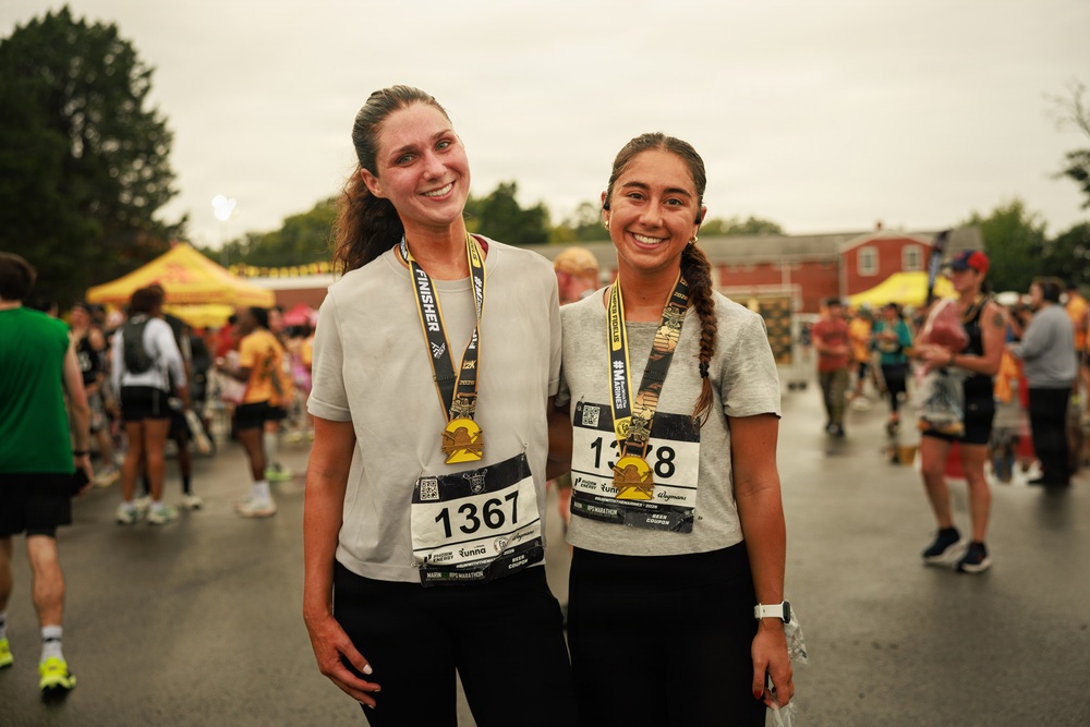Two smiling women pose together after completing a marathon race, both wearing finisher medals and race bibs. They are dressed in matching gray t-shirts and black pants, standing in a post-race festival area with tents and other participants visible in the background. The overcast, wet conditions suggest the race took place in rainy weather.