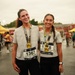 Two smiling women pose together after completing a marathon race, both wearing finisher medals and race bibs. They are dressed in matching gray t-shirts and black pants, standing in a post-race festival area with tents and other participants visible in the background. The overcast, wet conditions suggest the race took place in rainy weather.