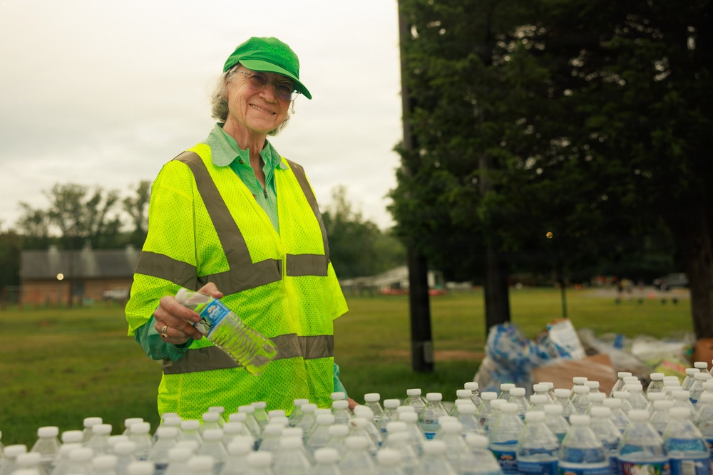 An older woman wearing a neon yellow high-visibility safety vest and green cap is smiling while distributing bottled water at what appears to be an outdoor event or volunteer operation. She is holding a water bottle in her hand and standing behind a table covered with numerous plastic water bottles. The setting is a grassy park or field with trees visible in the background under an overcast sky.