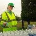 An older woman wearing a neon yellow high-visibility safety vest and green cap is smiling while distributing bottled water at what appears to be an outdoor event or volunteer operation. She is holding a water bottle in her hand and standing behind a table covered with numerous plastic water bottles. The setting is a grassy park or field with trees visible in the background under an overcast sky.