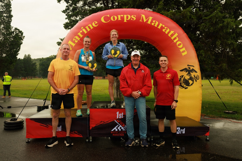 A podium ceremony at the Marine Corps Marathon, showing two female runners standing on 1st and 2nd place platforms beneath a large inflatable arch bearing the event name. Both athletes are holding heart-shaped awards/trophies. Four additional individuals, including what appear to be race officials or staff, stand alongside the podium.