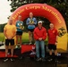 A podium ceremony at the Marine Corps Marathon, showing two female runners standing on 1st and 2nd place platforms beneath a large inflatable arch bearing the event name. Both athletes are holding heart-shaped awards/trophies. Four additional individuals, including what appear to be race officials or staff, stand alongside the podium.