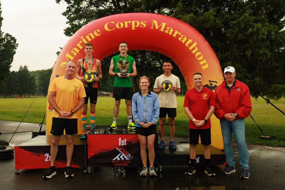 A podium ceremony at the Marine Corps Marathon featuring winners standing on 1st, 2nd, and 3rd place platforms beneath an inflatable arch bearing the event name. Several participants are wearing medals and holding trophies, accompanied by what appear to be race officials or staff. The event is taking place outdoors in wet, overcast conditions.