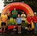 A podium ceremony at the Marine Corps Marathon featuring winners standing on 1st, 2nd, and 3rd place platforms beneath an inflatable arch bearing the event name. Several participants are wearing medals and holding trophies, accompanied by what appear to be race officials or staff. The event is taking place outdoors in wet, overcast conditions.