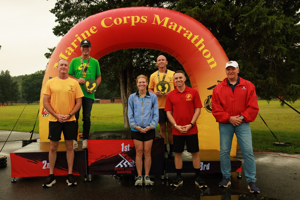 A group of seven people pose at a Marine Corps Marathon awards ceremony in front of an inflatable arch bearing the event name. Two athletes stand elevated on a podium marked with 1st, 2nd, and 3rd place positions, with at least two individuals holding trophy plaques and wearing medals. The setting appears to be outdoors on a wet, overcast day.