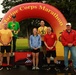 A group of seven people pose at a Marine Corps Marathon awards ceremony in front of an inflatable arch bearing the event name. Two athletes stand elevated on a podium marked with 1st, 2nd, and 3rd place positions, with at least two individuals holding trophy plaques and wearing medals. The setting appears to be outdoors on a wet, overcast day.