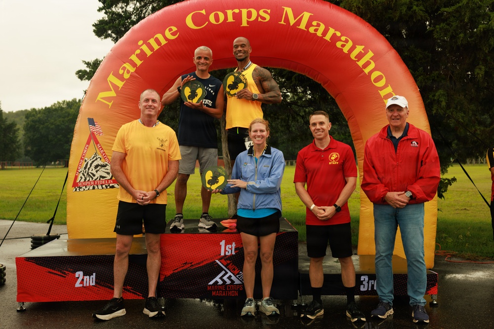 A podium ceremony at the Marine Corps Marathon event, showing winners standing on 1st, 2nd, and 3rd place platforms beneath a large inflatable arch bearing the event name. Two athletes in yellow running gear stand elevated on the podium holding trophy awards, while a woman in blue holds a trophy on the 1st place platform. Several officials or staff members in red and yellow shirts flank the podium.