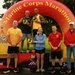 A podium ceremony at the Marine Corps Marathon event, showing winners standing on 1st, 2nd, and 3rd place platforms beneath a large inflatable arch bearing the event name. Two athletes in yellow running gear stand elevated on the podium holding trophy awards, while a woman in blue holds a trophy on the 1st place platform. Several officials or staff members in red and yellow shirts flank the podium.