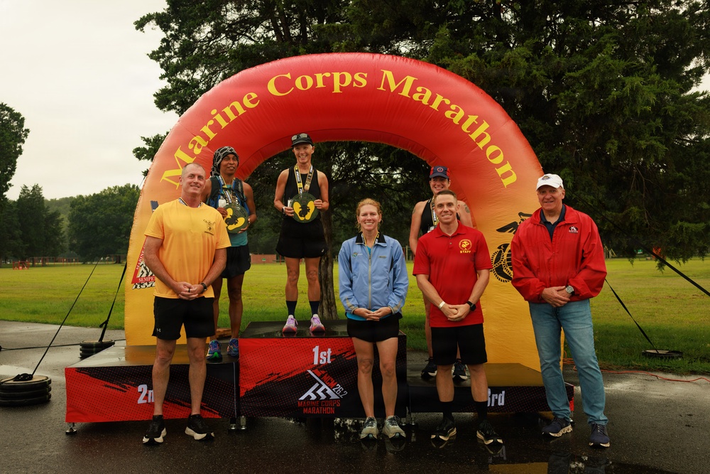 A podium ceremony at the Marine Corps Marathon featuring three place finishers standing on 1st, 2nd, and 3rd place platforms beneath an inflatable arch. The event appears to be taking place in wet, overcast conditions on a paved surface surrounded by green grass and trees. Several officials or staff members flank the podium alongside the medal-wearing competitors.